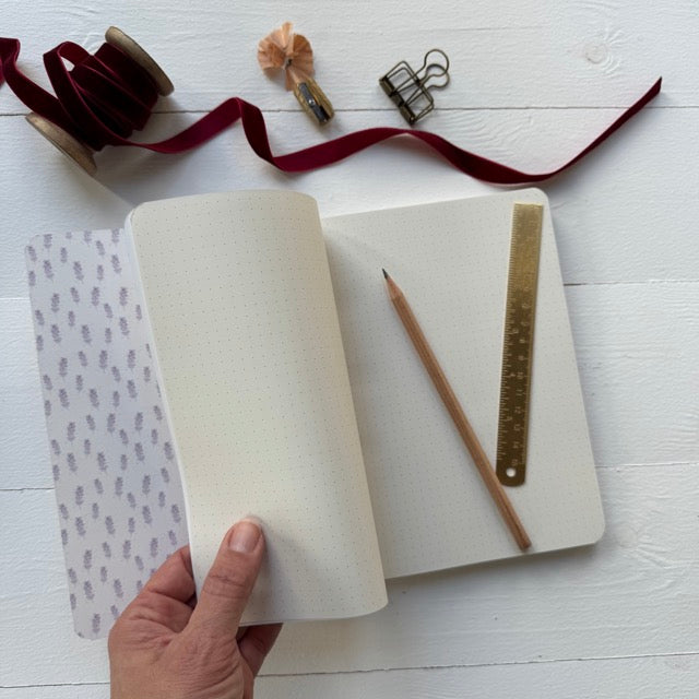 Notebook being held open by a hand with pen and ruler on a white surface with decorative items.