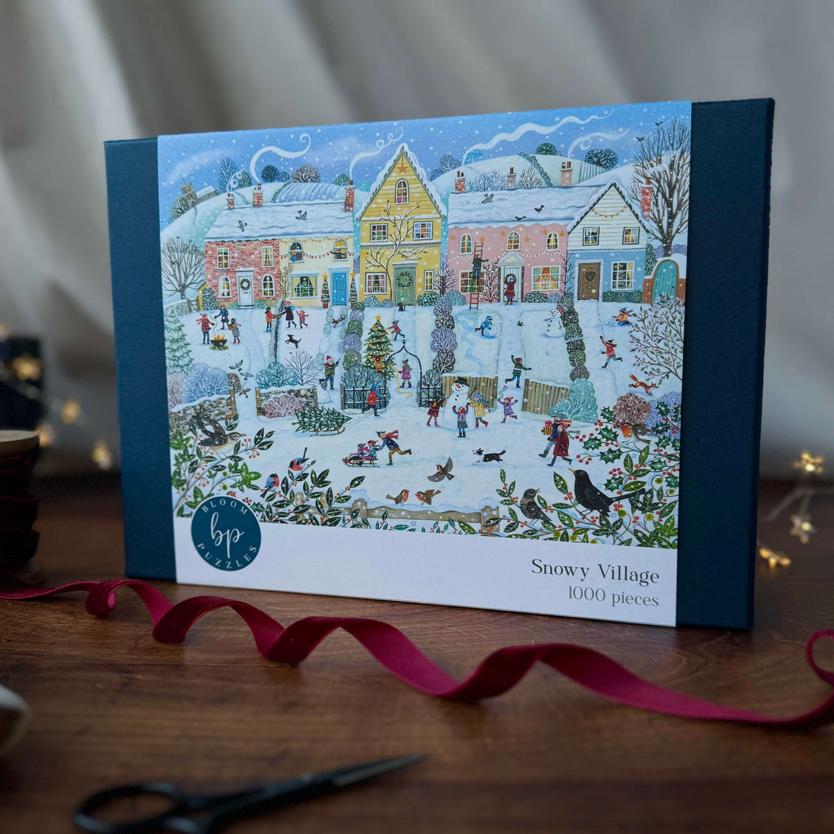 A boxed jigsaw puzzle titled "Snowy Village" stands on a wooden table. The front of the box shows a detailed winter landscape illustration with a row of coloured houses, people enjoying sledging and outdoor activities in the snow.