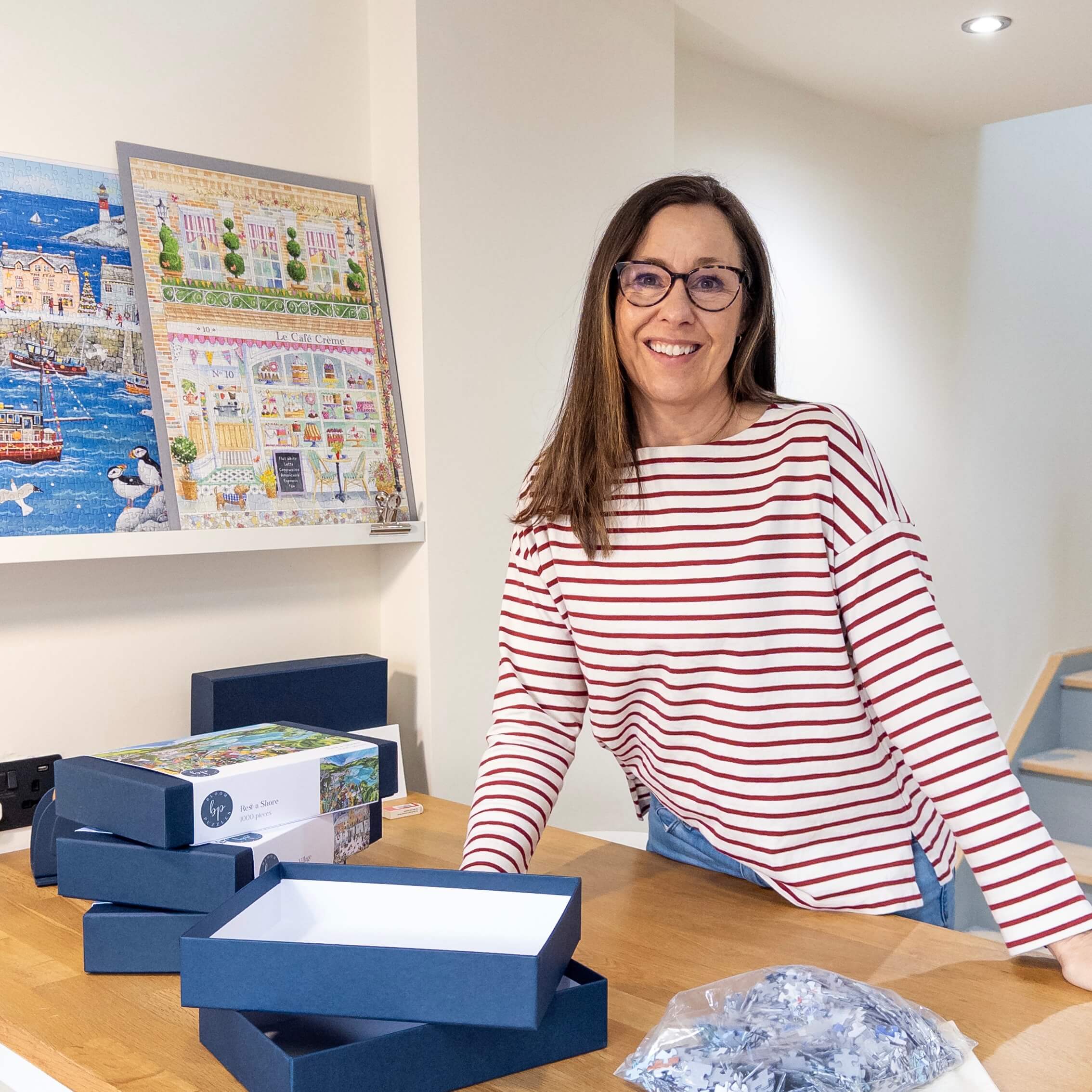 Philippa in a striped top standing behind the packing table with puzzle boxes and a puzzles.