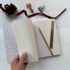 Notebook being held open by a hand with pen and ruler on a white surface with decorative items.
